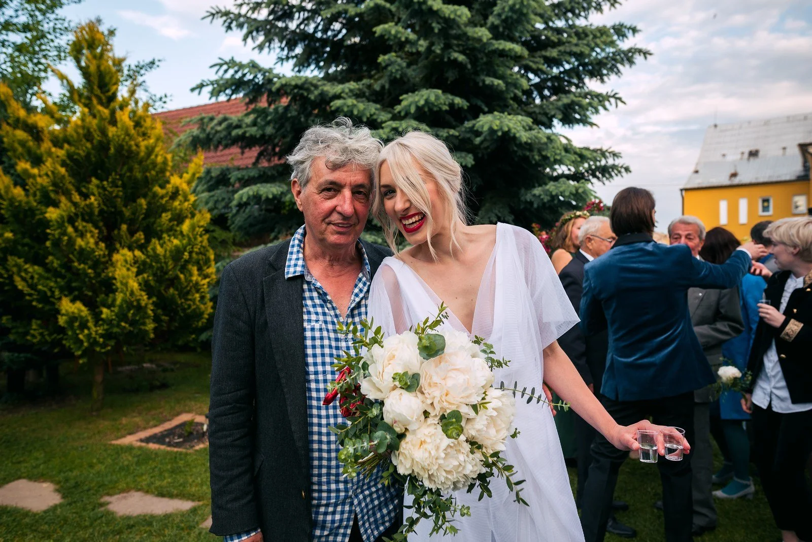 Žena v bílých šatech s kyticí stojí v zahradě těsně vedle staršího muže.