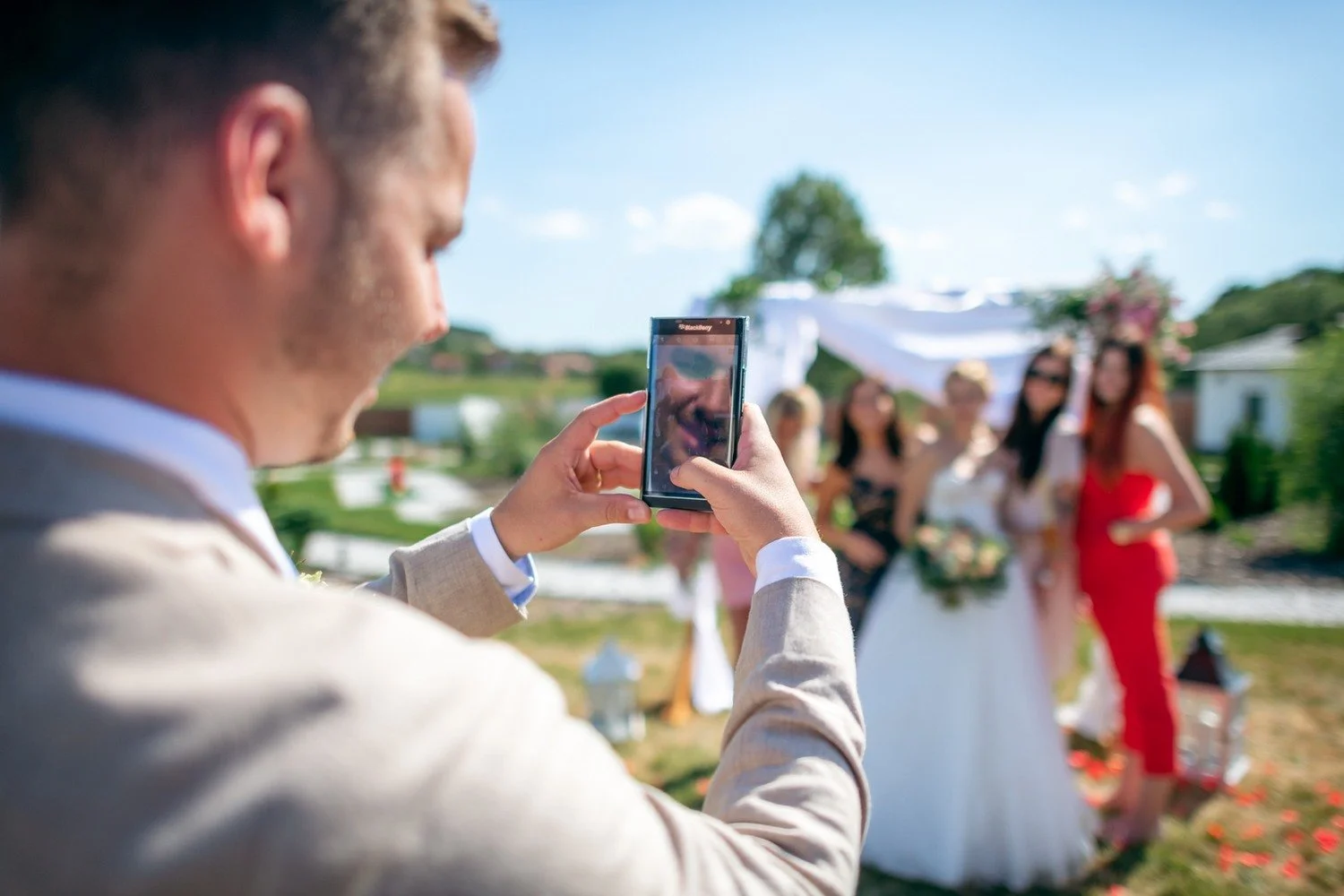 Muž v obleku fotografuje telefonem skupinu žen pózujících venku u květinové výzdoby.