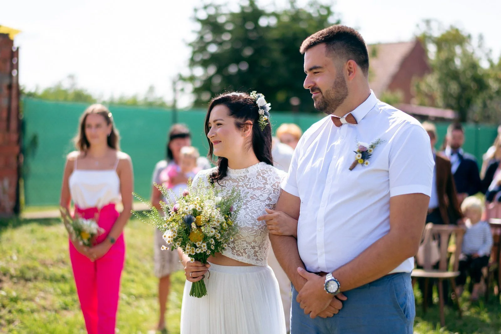 Žena s kyticí a muž s motýlkem stojí vedle sebe venku před rozostřenými hosty.