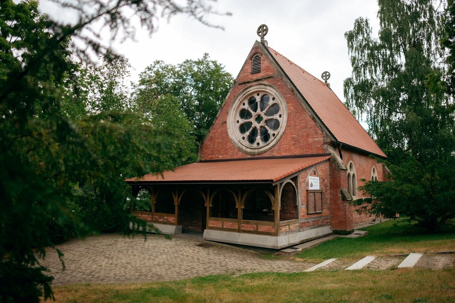 Cihlový kostel s kruhovým oknem a krytou vstupní verandou obklopují vzrostlé stromy.