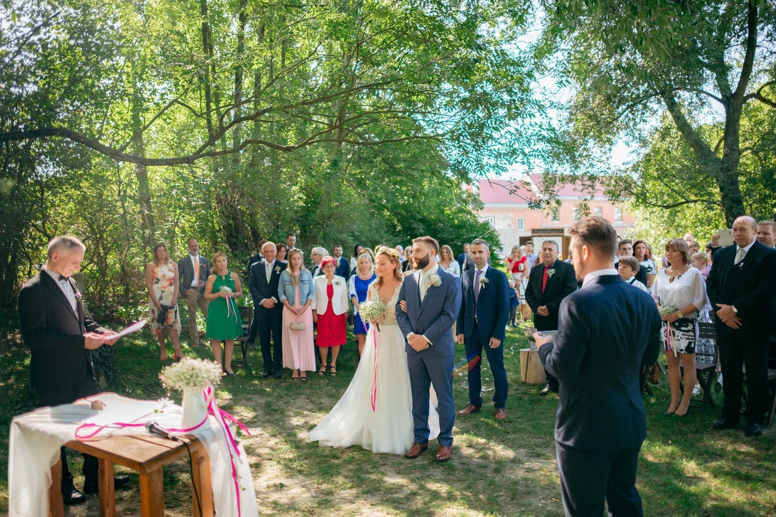 Široký pohled zachycuje venkovní obřad pod stromy s hosty rozestavenými kolem dvojice.