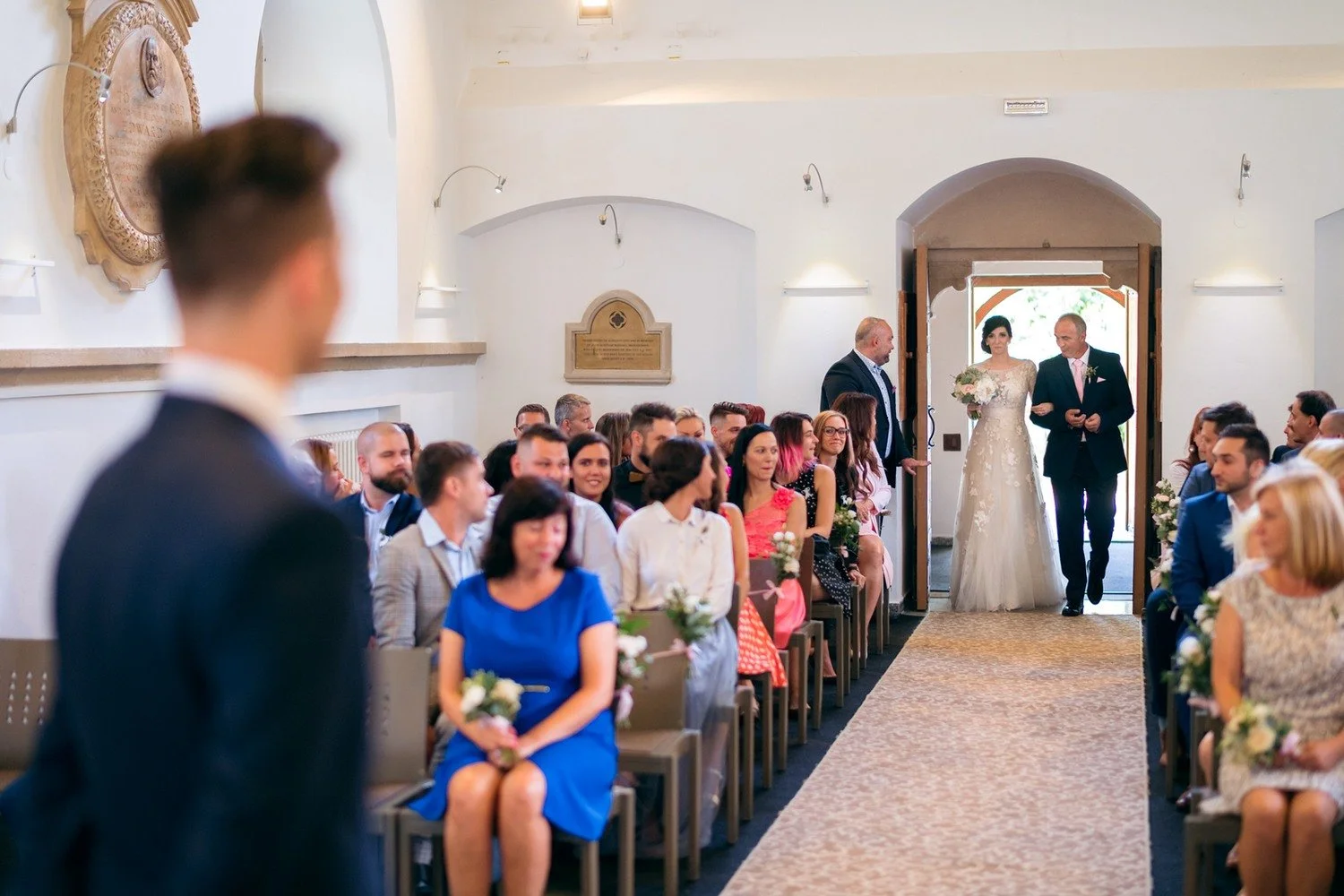 Nevěsta přichází středem zaplněného kostela po boku staršího muže, zatímco ženich čeká v popředí.