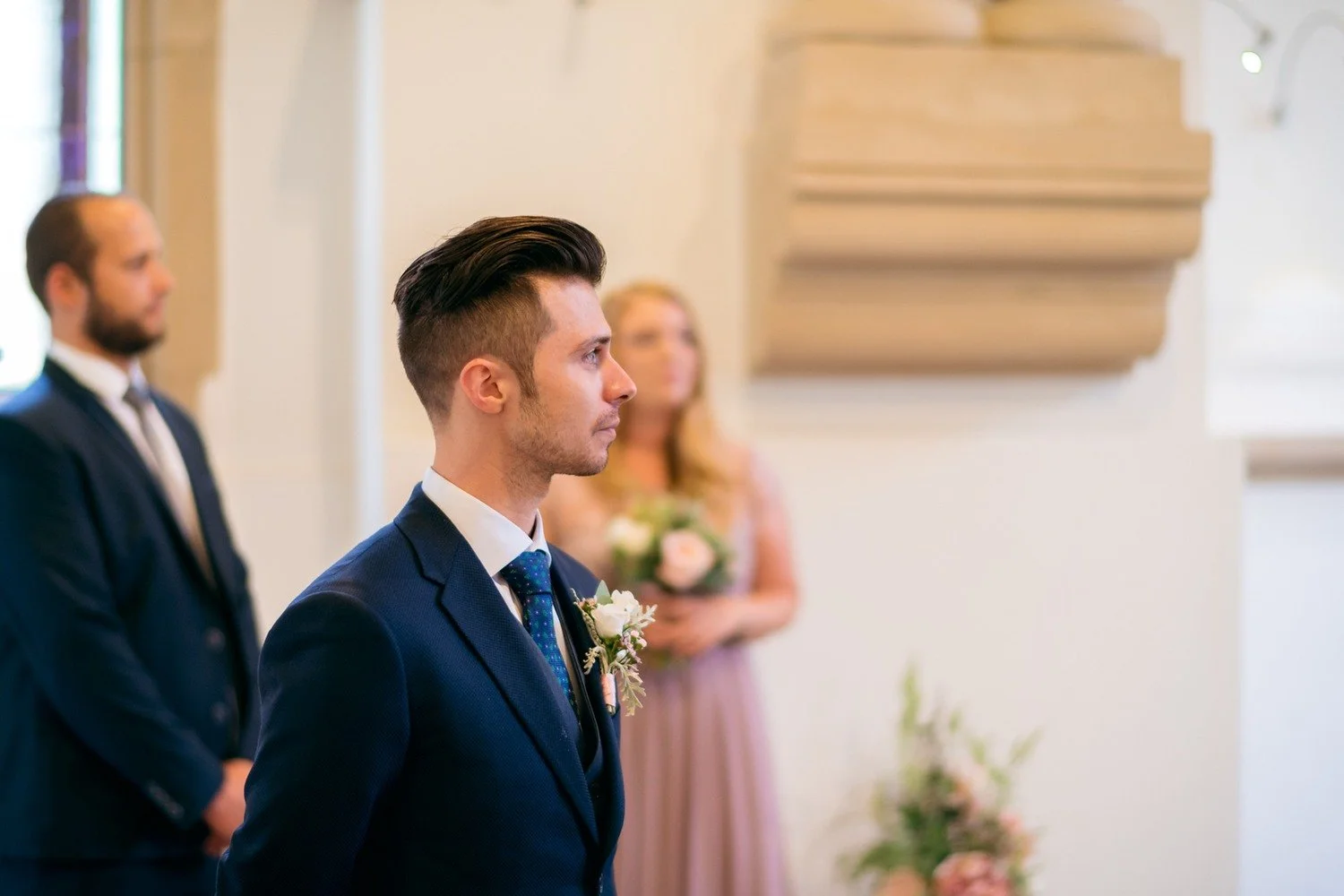 Vašek v tmavomodrém obleku stojí v profilu uvnitř kostela, za ním jsou rozostřené dvě osoby.