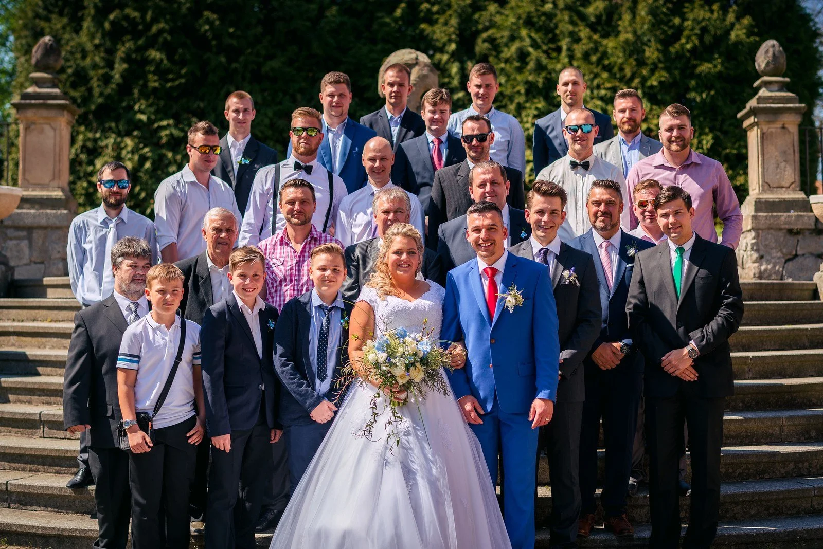 Skupinová fotografie mužů s novomanželi venku po obřadu.