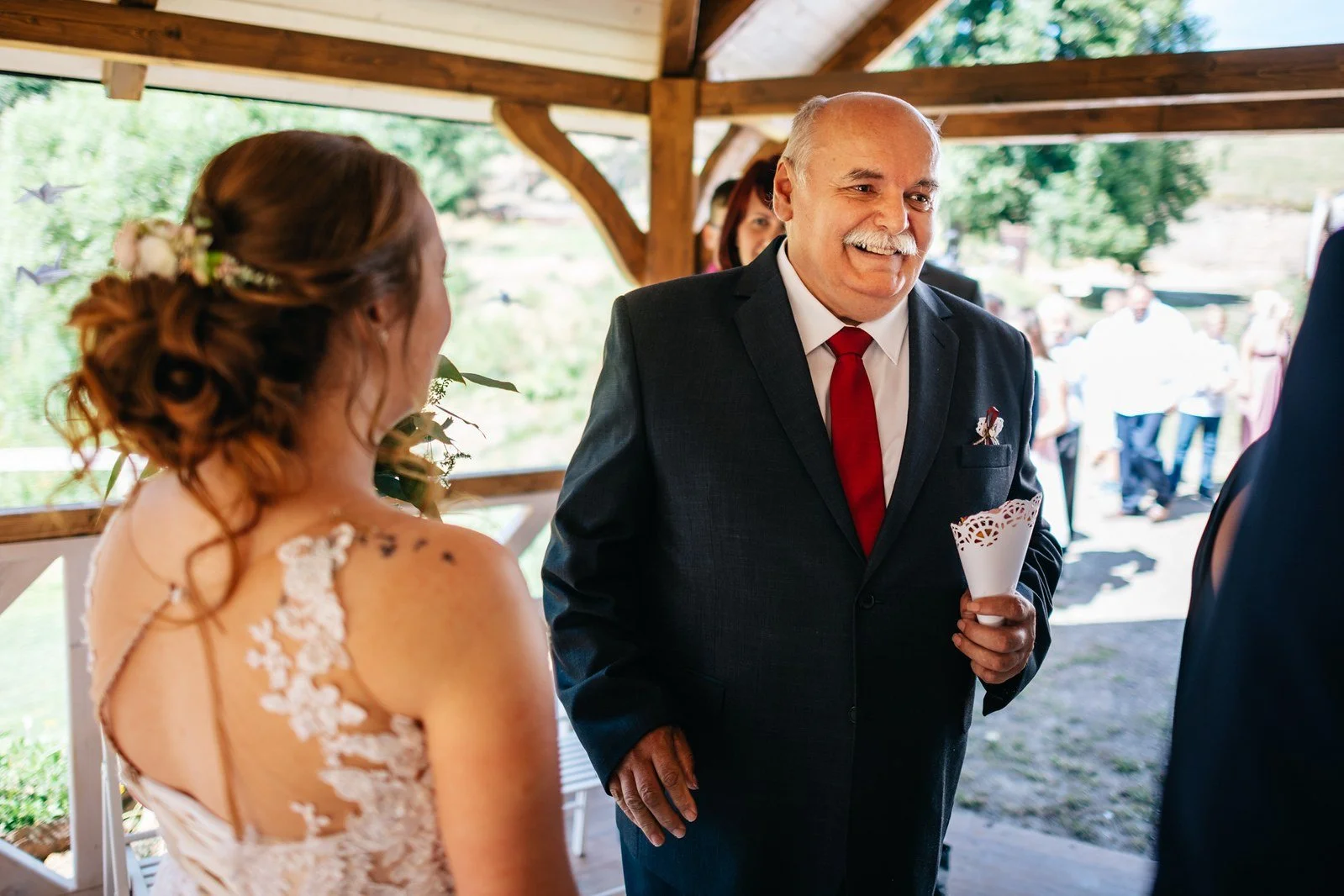 Hosté sedí na židlích při venkovním obřadu pod vzrostlými stromy.