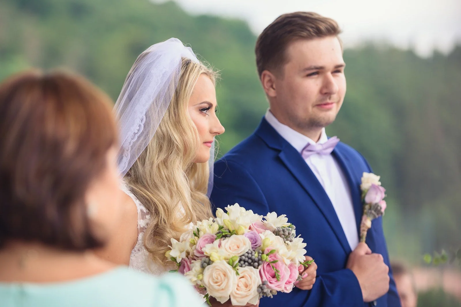 Nevěsta s kyticí a ženich si hledí do očí během venkovního obřadu.