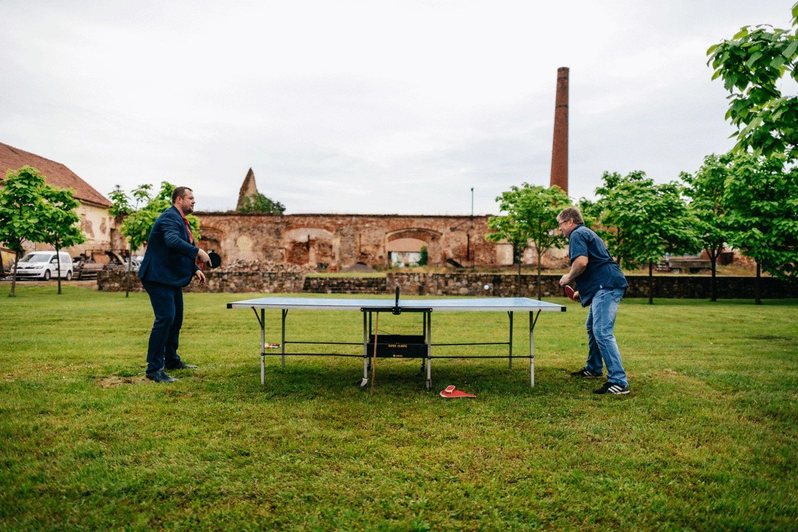 Dva lidé hrají stolní tenis na trávníku, za nimiž se táhne nízká kamenná stavba a vysoký komín.