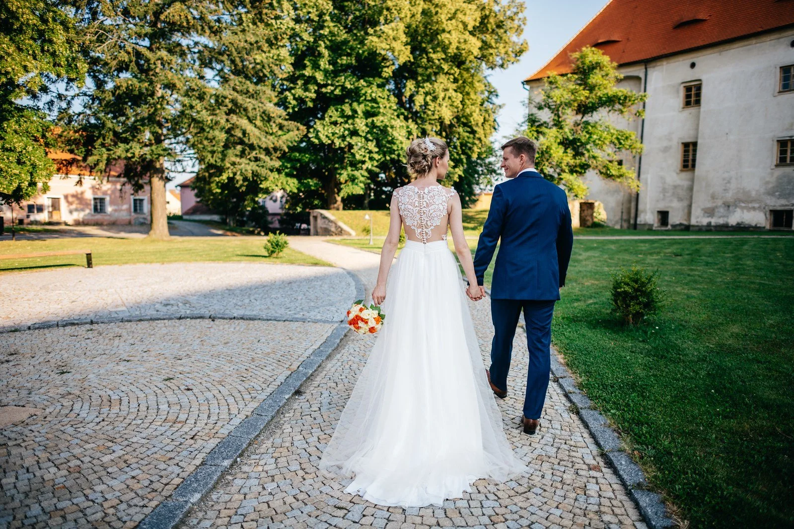 Novomanželé odcházejí po kamenné cestě mezi budovami.