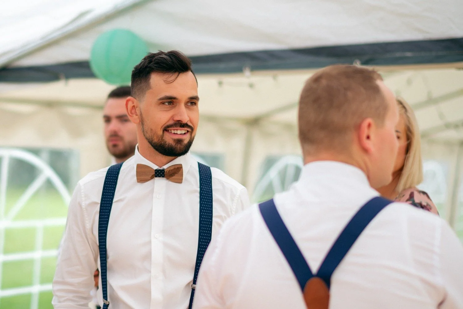Ženich s dřevěným motýlkem a modrými kšandami se usmívá při rozhovoru pod stanem.
