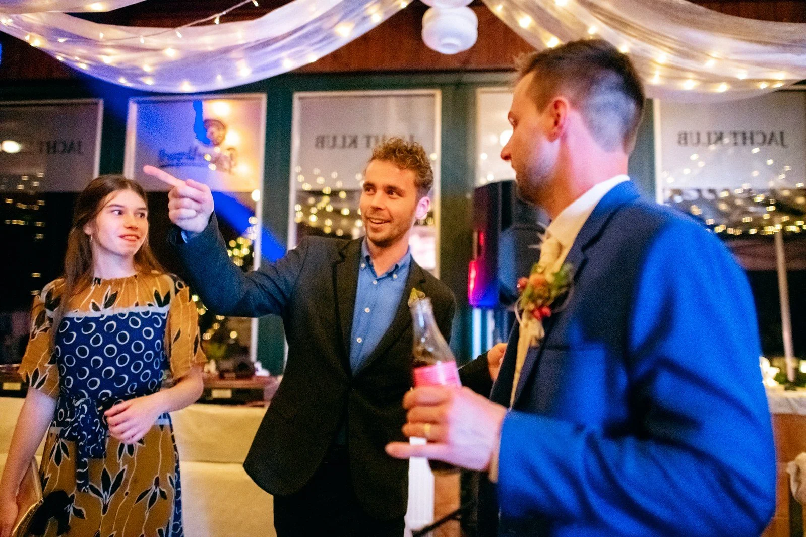 Muž s lahví stojí v popředí, zatímco další muž ukazuje prstem přes místnost.