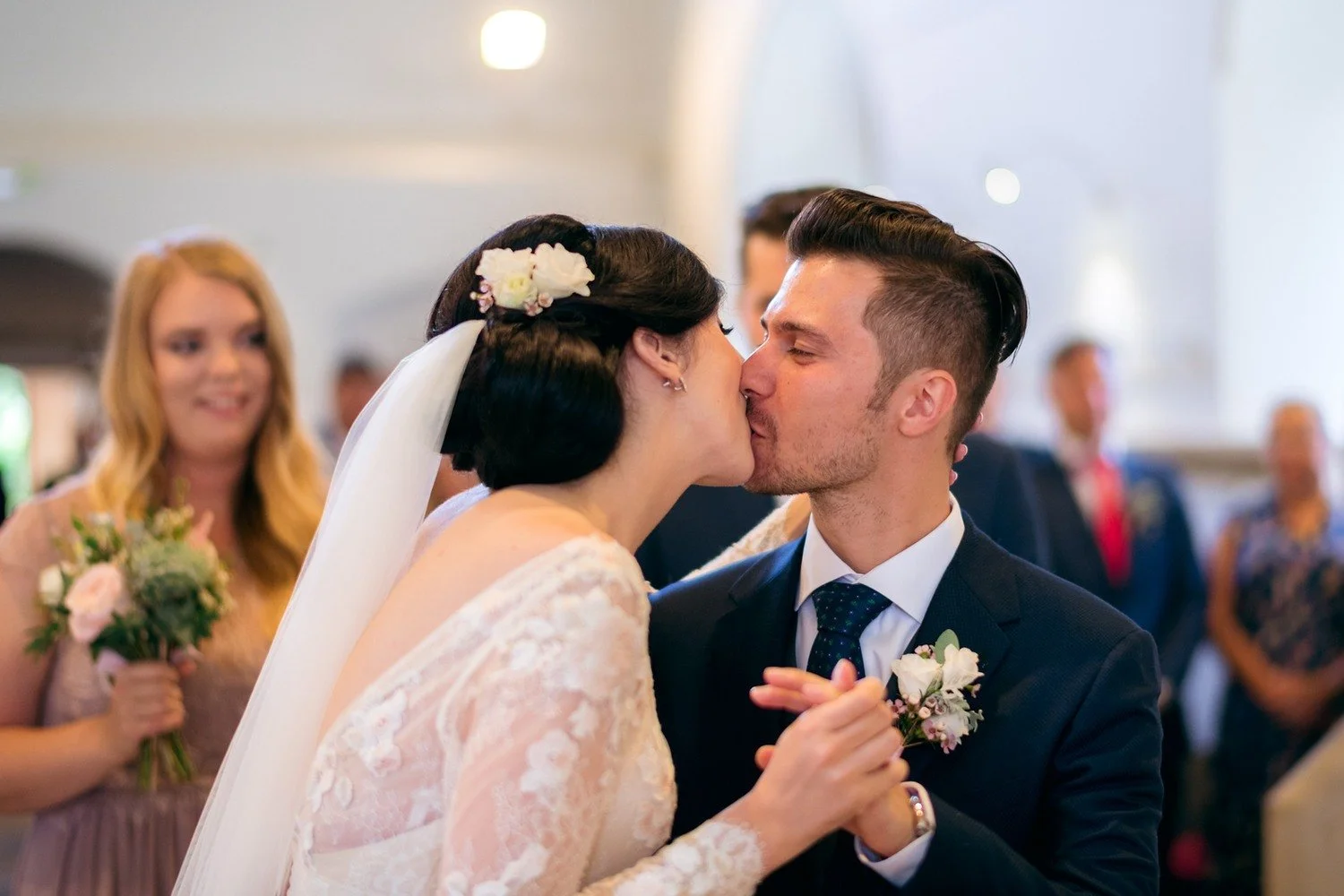 Kateřina a Vašek se líbají a drží se za ruce, zatímco žena s kyticí stojí rozostřená v pozadí.