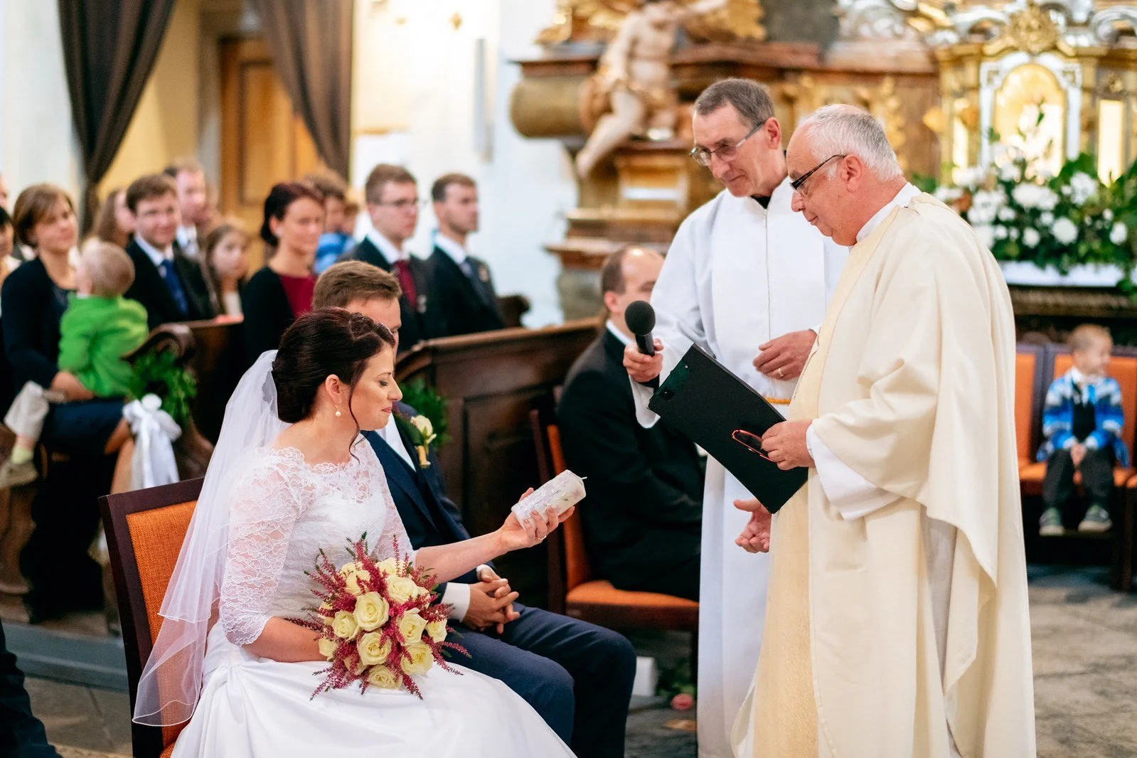 Sedící žena v bílých šatech drží svíci, zatímco před ní stojí dva muži ve světlých liturgických oděvech.