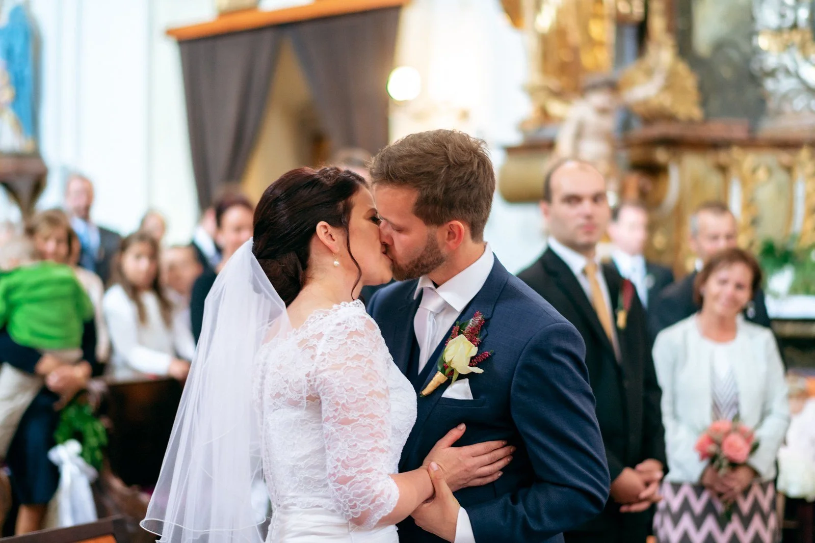 Dvojice ve svatebním oblečení se líbá před oltářem a řadami přihlížejících.