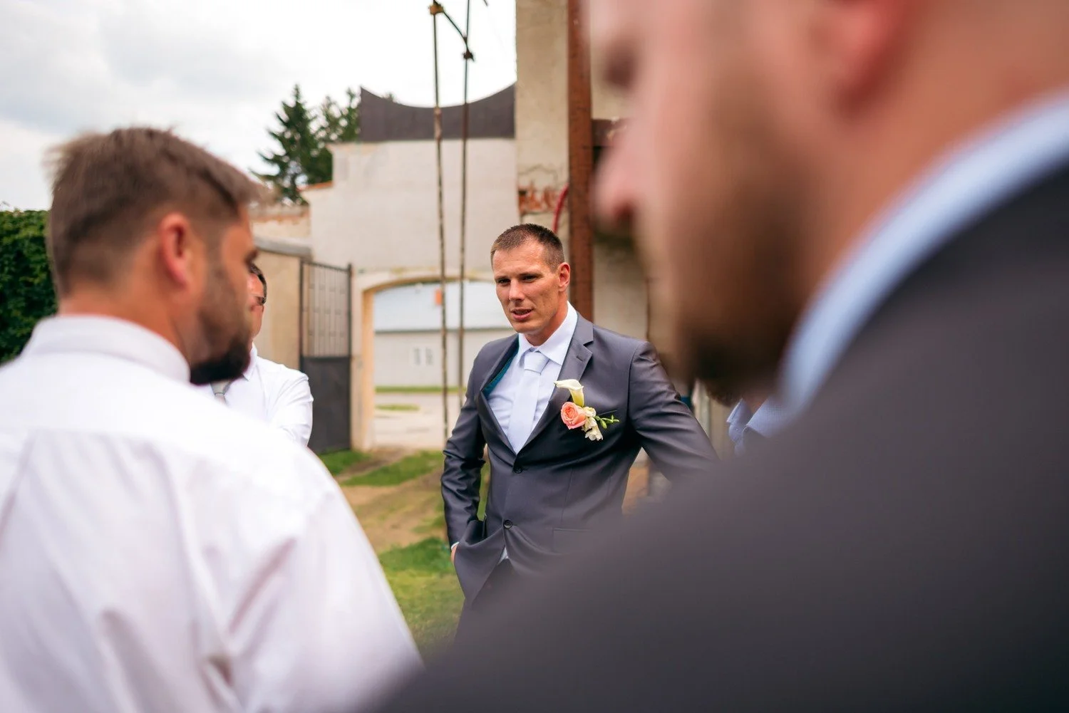 Lukáš v tmavém obleku stojí venku mezi dalšími muži, jejichž rozostřená ramena rámují záběr.