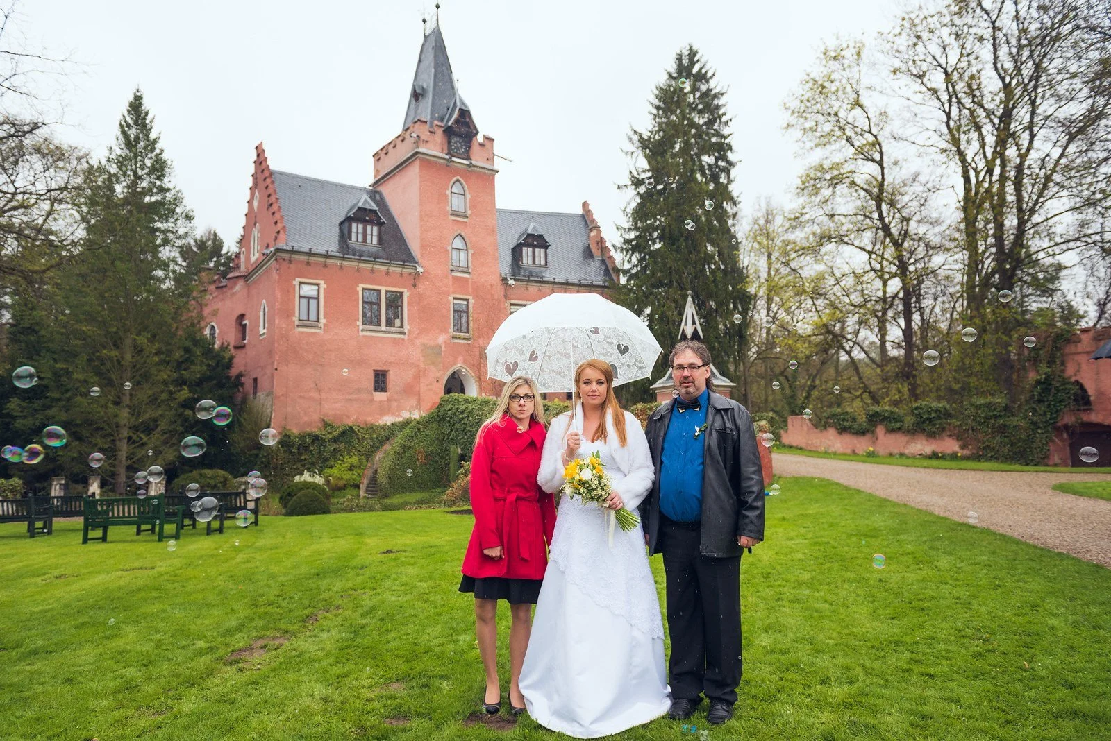 Nevěsta se ženichem stojí pod průhledným deštníkem v zámeckém parku.