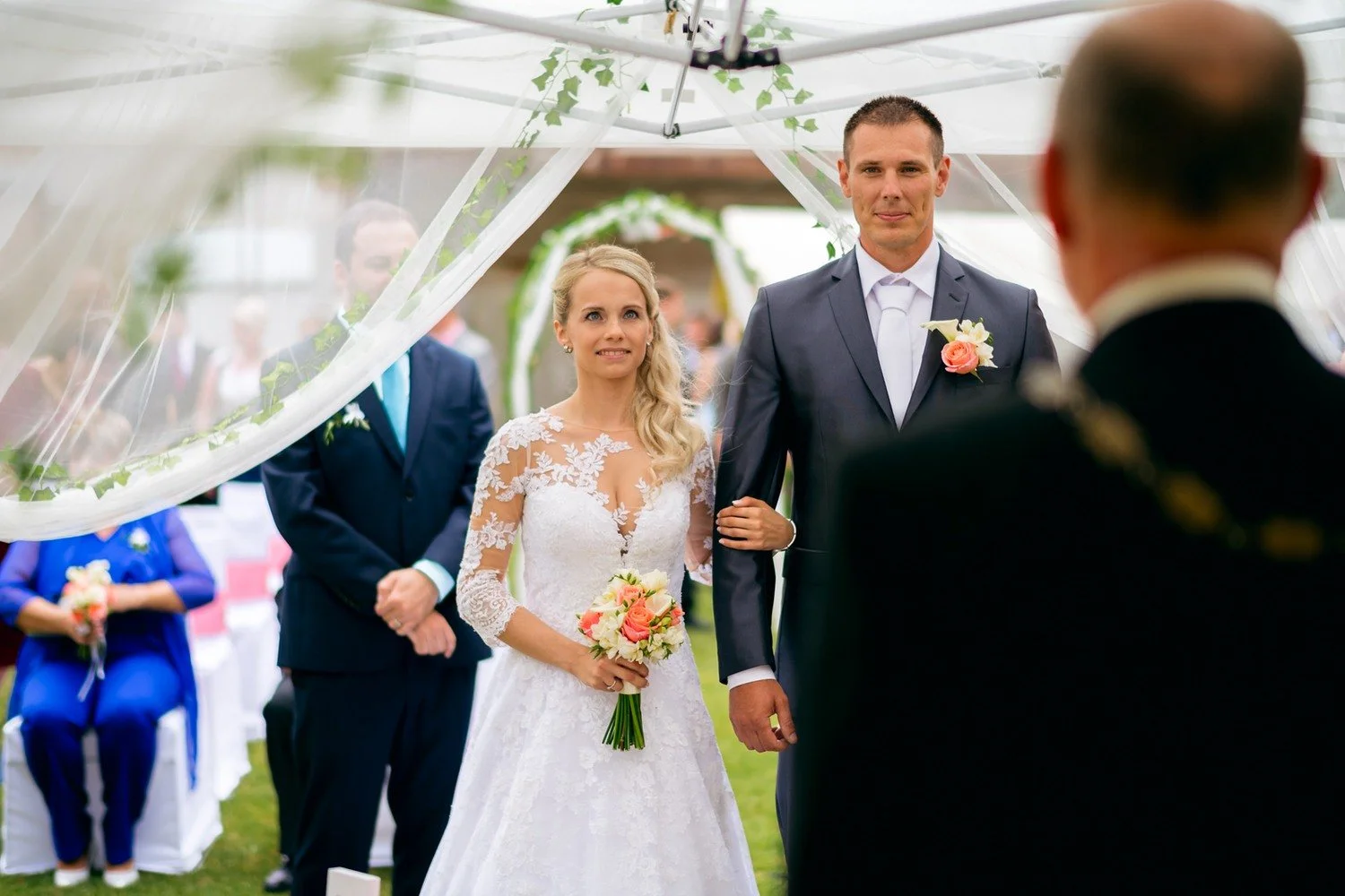 Nevěsta přichází pod průsvitný stan zavěšená do vysokého muže, zatímco v popředí stojí další muž zády.