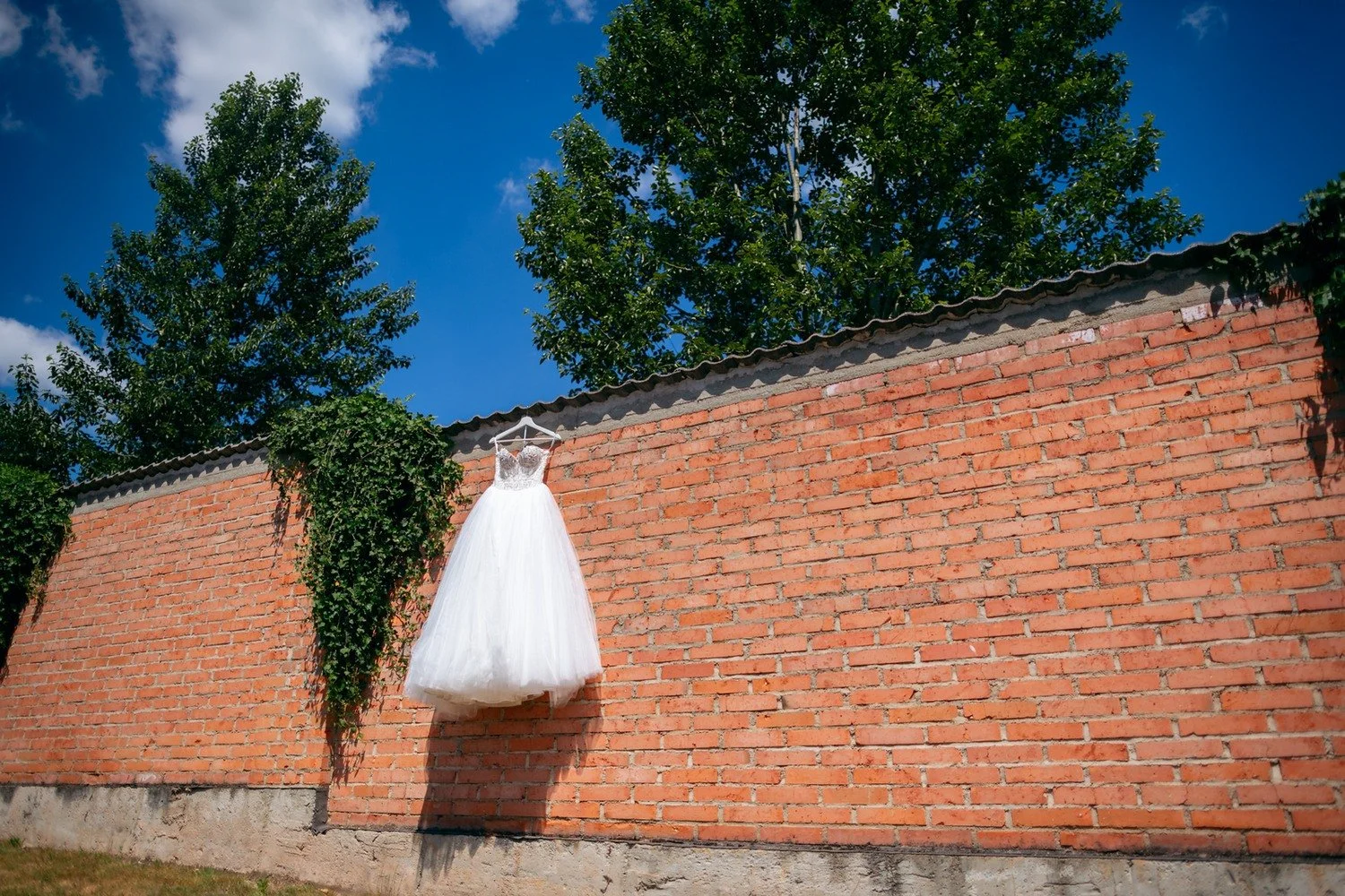 Bílé svatební šaty zavěšené venku na vysoké cihlové zdi.