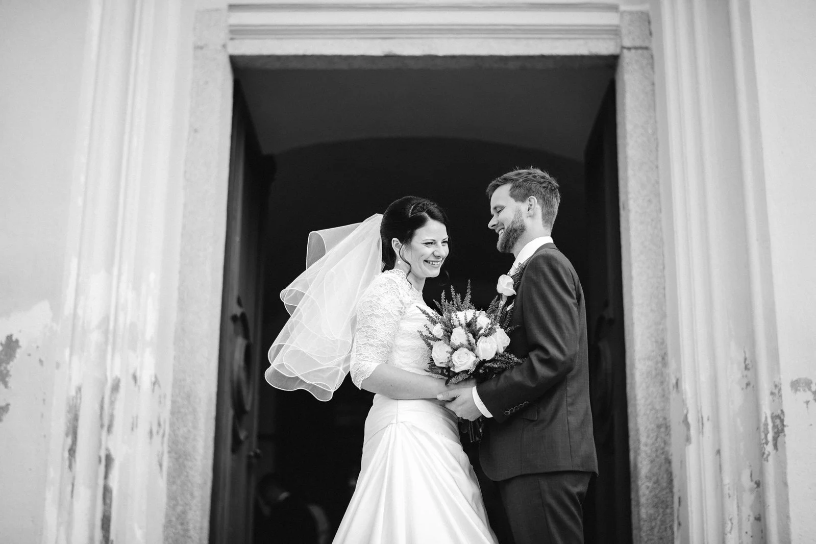 Černobílá fotografie rámuje dvojici ve svatebním oblečení v otevřených dveřích kostela.