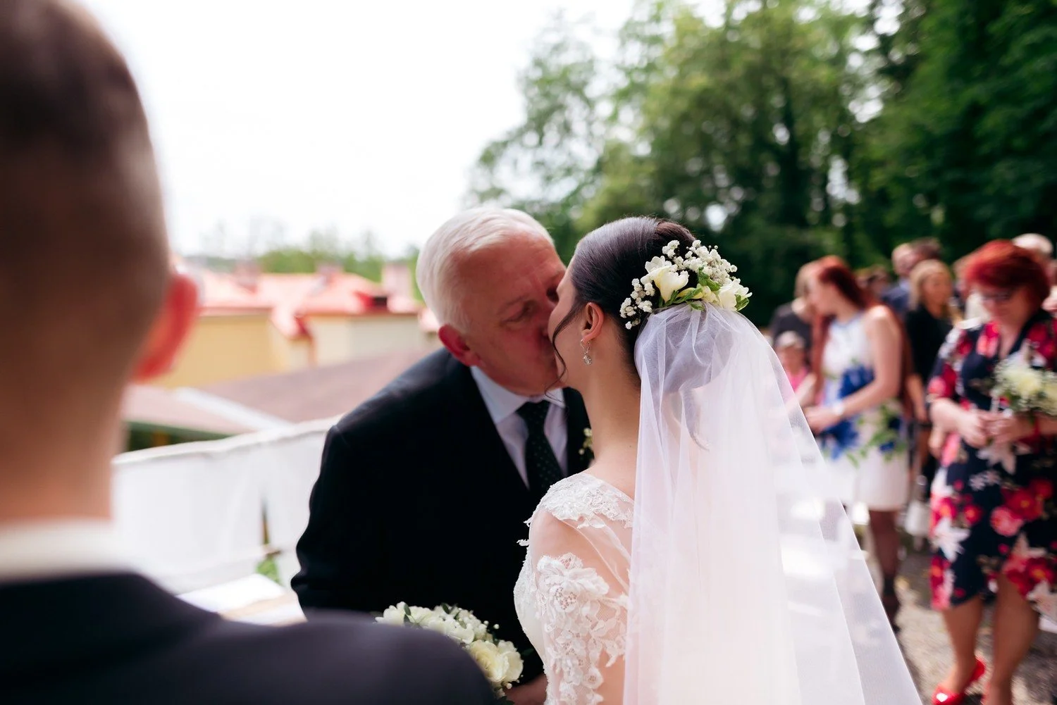 Starší muž líbá nevěstu na tvář mezi ostatními gratulujícími.