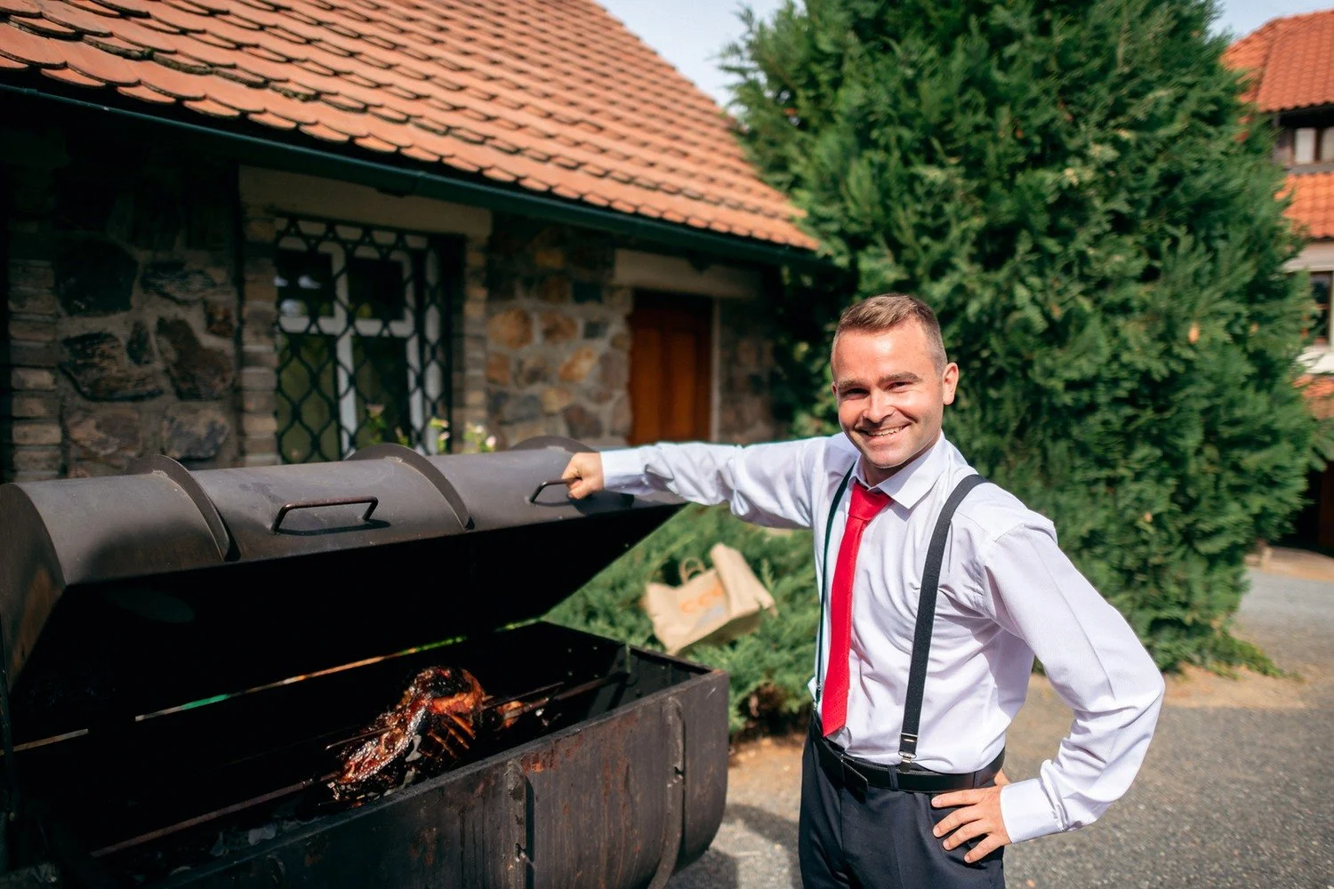 Usměvavý muž v bílé košili obrací maso na velkém venkovním grilu.