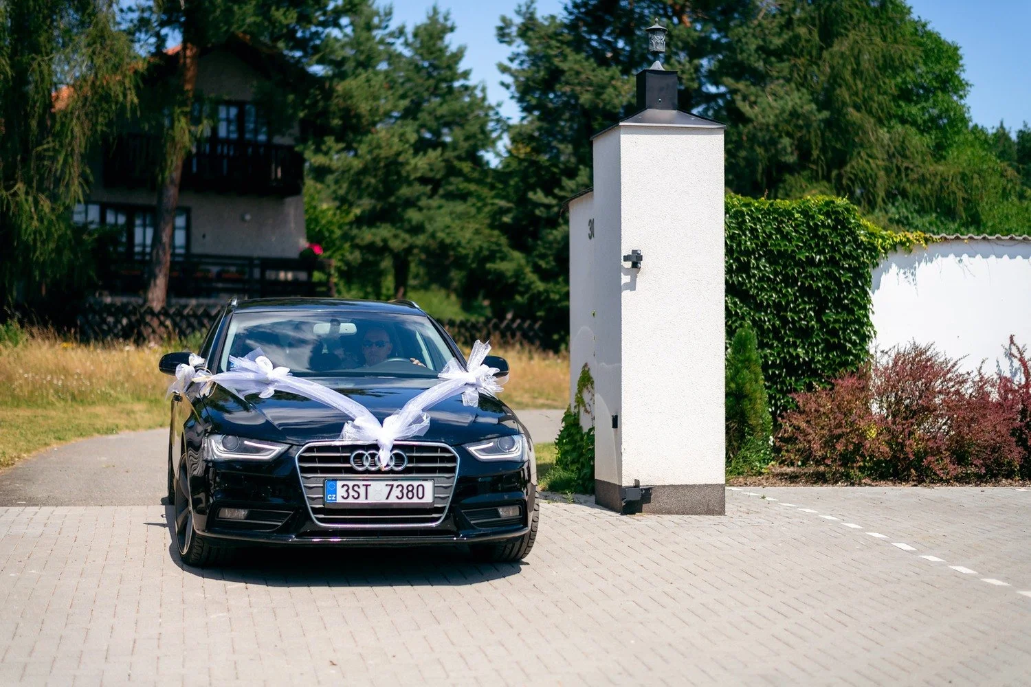 Černý automobil ozdobený bílými stuhami přijíždí otevřenou bránou.