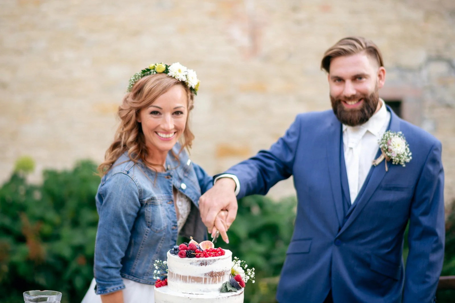 Žena s květinovým věnečkem a muž v modrém obleku společně krájejí dort u venkovního stolu.