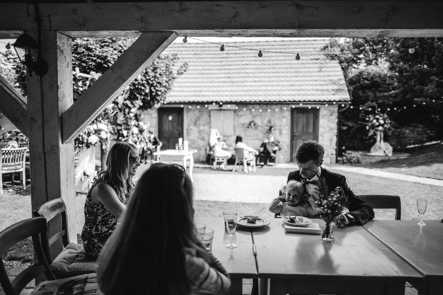 Černobílý pohled z tmavého přístřešku směrem ke stolům a lidem na slunném dvoře.