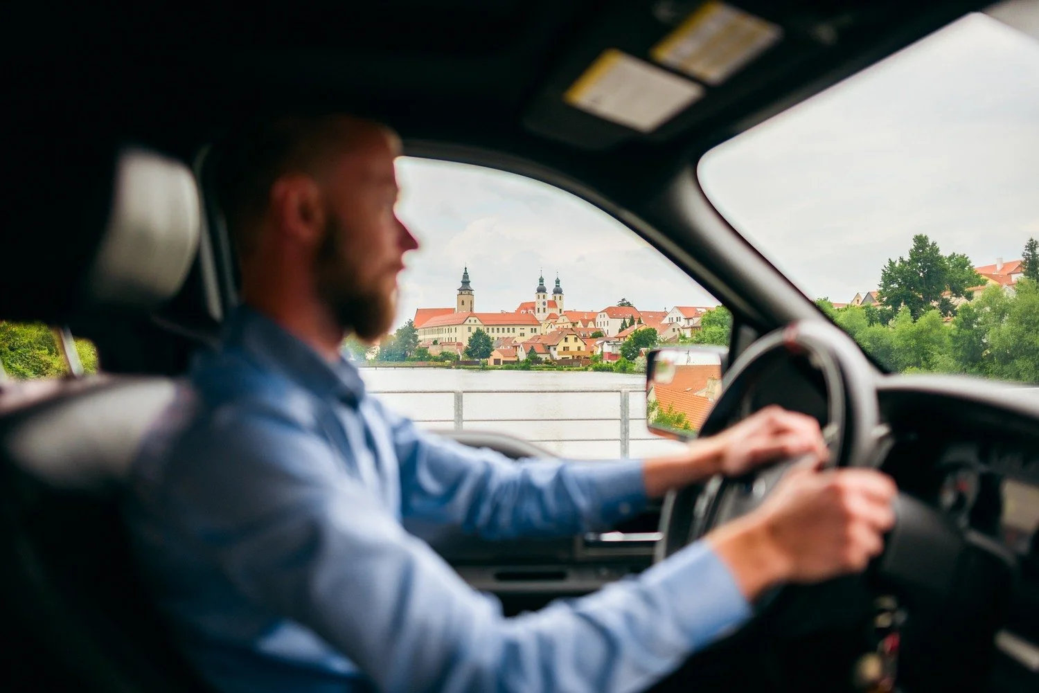 Řidič zachycený z profilu vede auto a za bočním oknem se rýsuje panorama města.