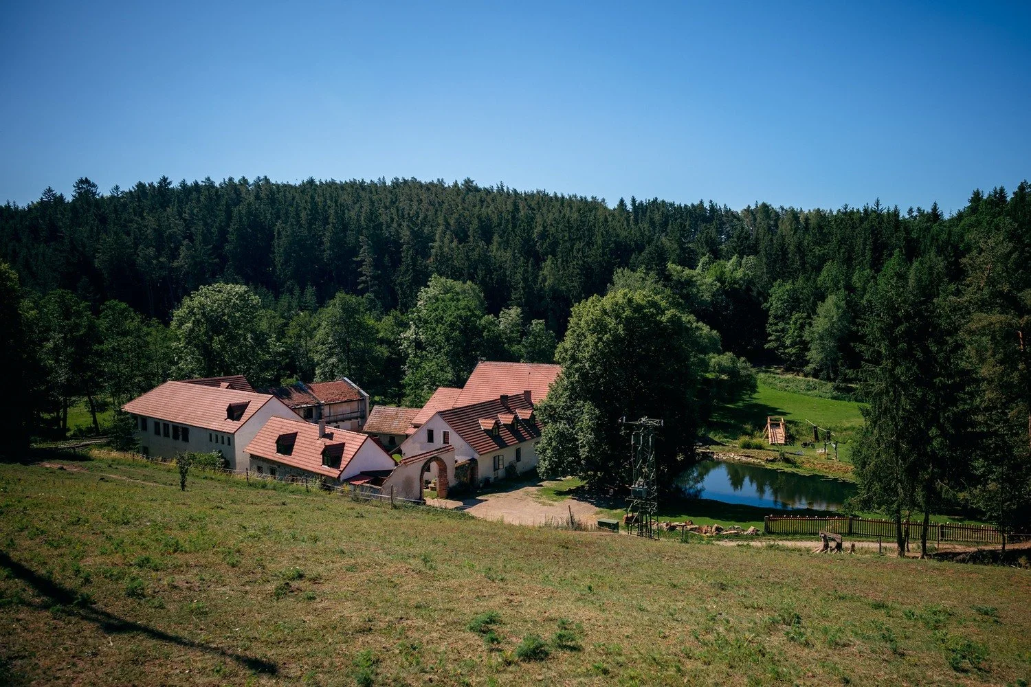 Letecký pohled ukazuje venkovský areál obklopený loukami, lesem a malým rybníkem.