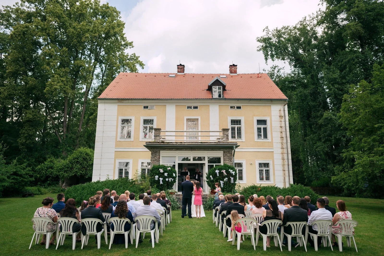 Venkovní obřad probíhá na trávníku před historickou budovou.