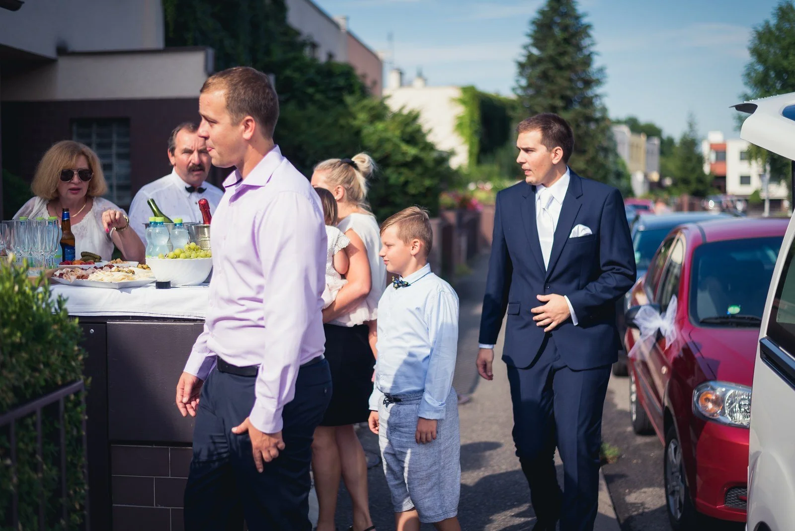 Skupina formálně oblečených lidí kráčí mezi zaparkovanými auty a posouvá děj ven.