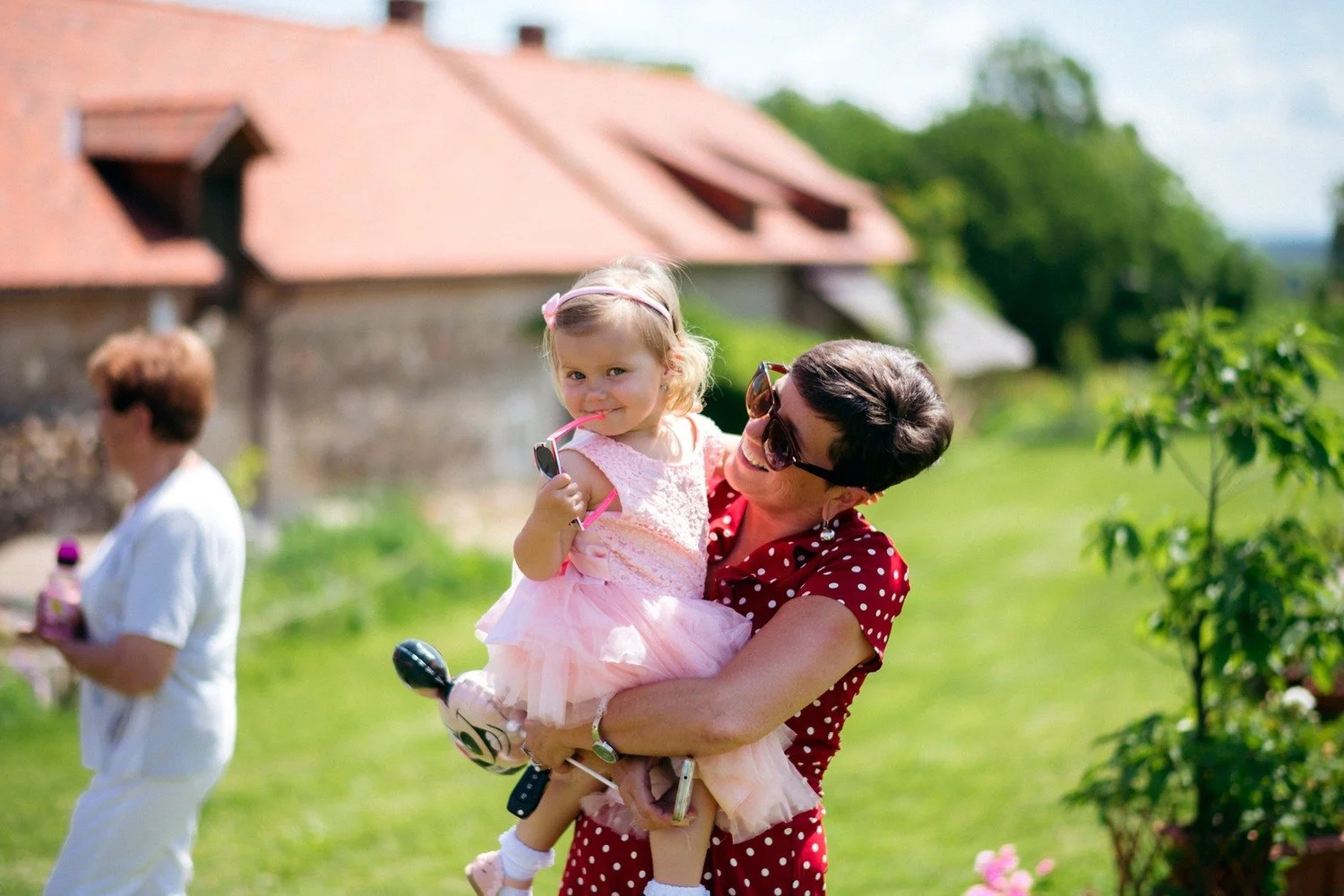 Žena v červených puntíkovaných šatech drží holčičku v růžových šatech a slunečních brýlích.