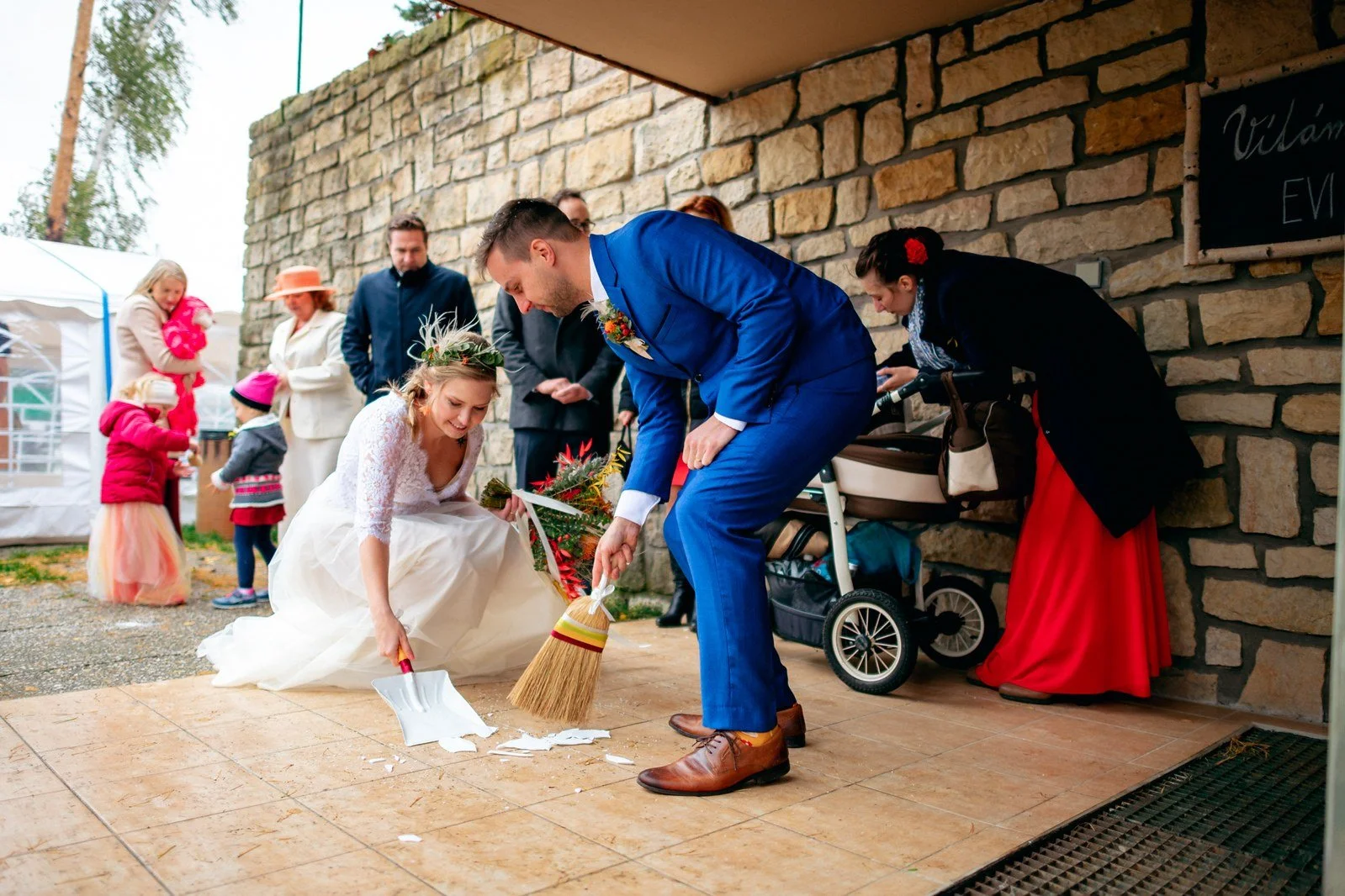 Muž v modrém obleku drží koště vedle přikrčené ženy v bílých šatech na dlážděné terase.