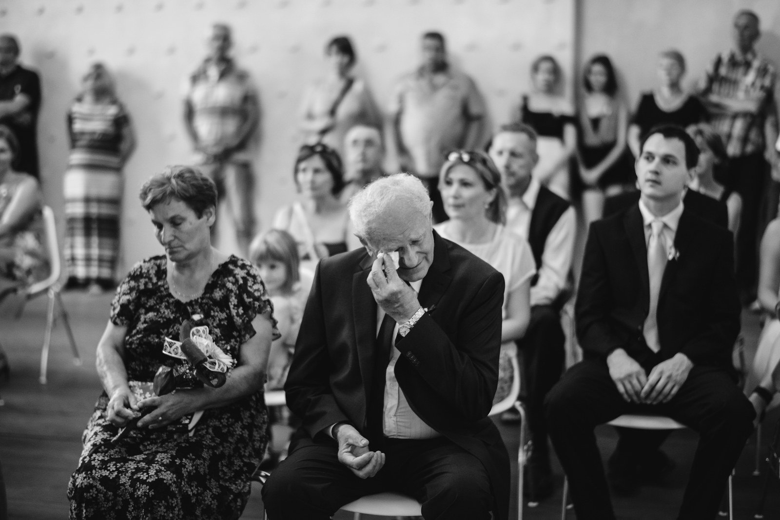 Černobílý snímek zachycuje pozorné tváře hostů během obřadu.