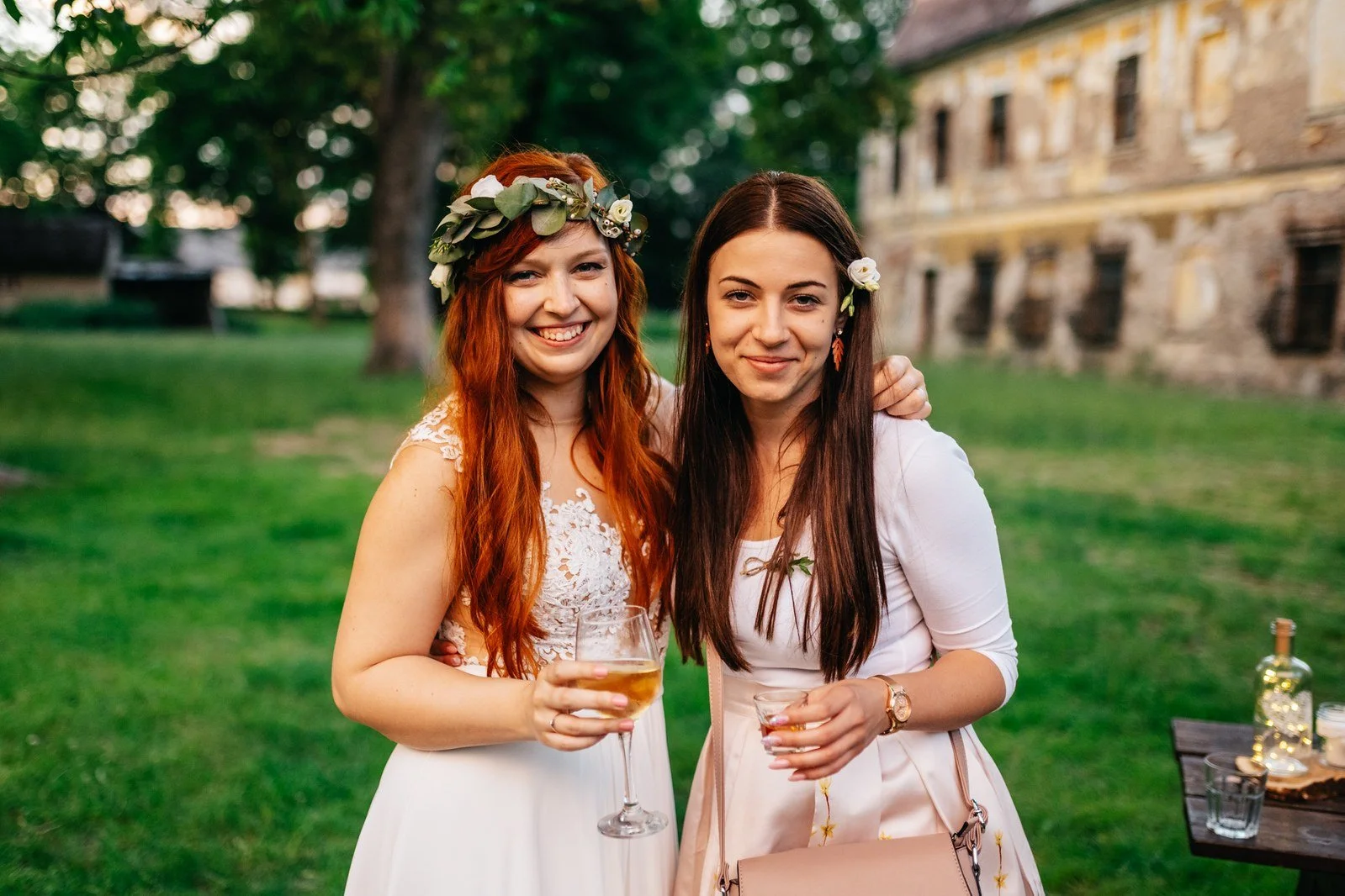 Dvě ženy s dlouhými vlasy pózují s nápoji v zahradě.
