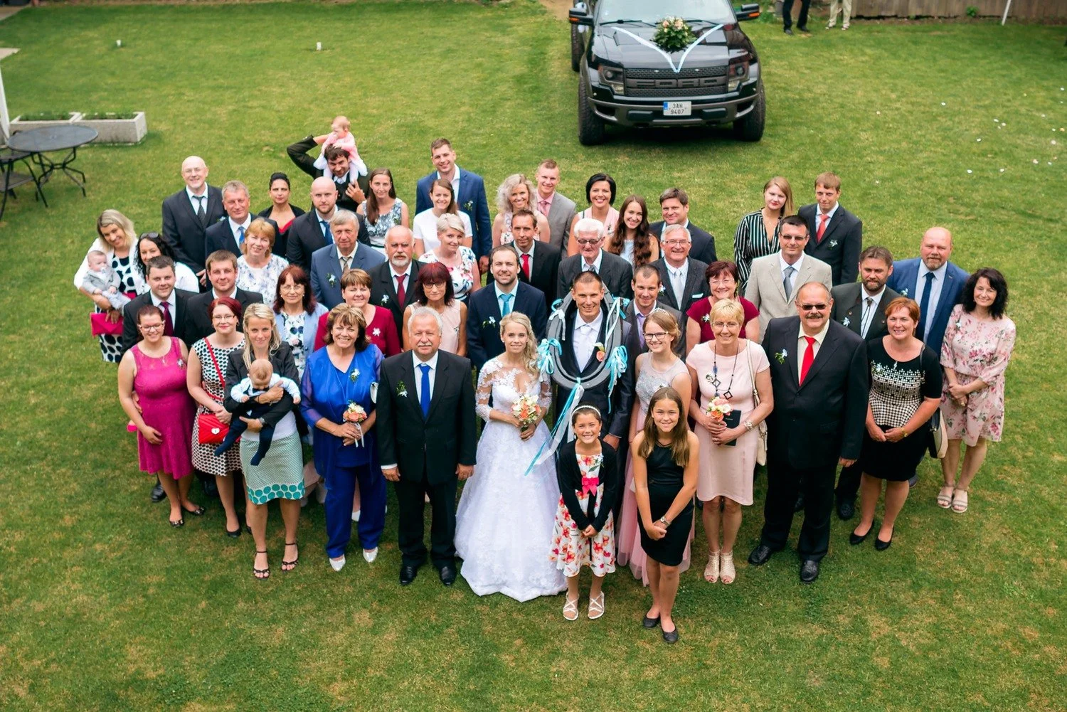 Velká skupina svatebních hostů se dívá vzhůru k fotoaparátu z vyvýšeného pohledu.