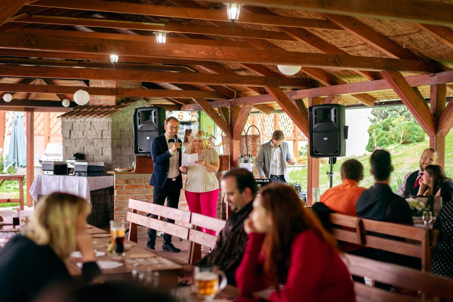 Muž pronáší řeč k sedícím hostům v dřevěném pavilonu.