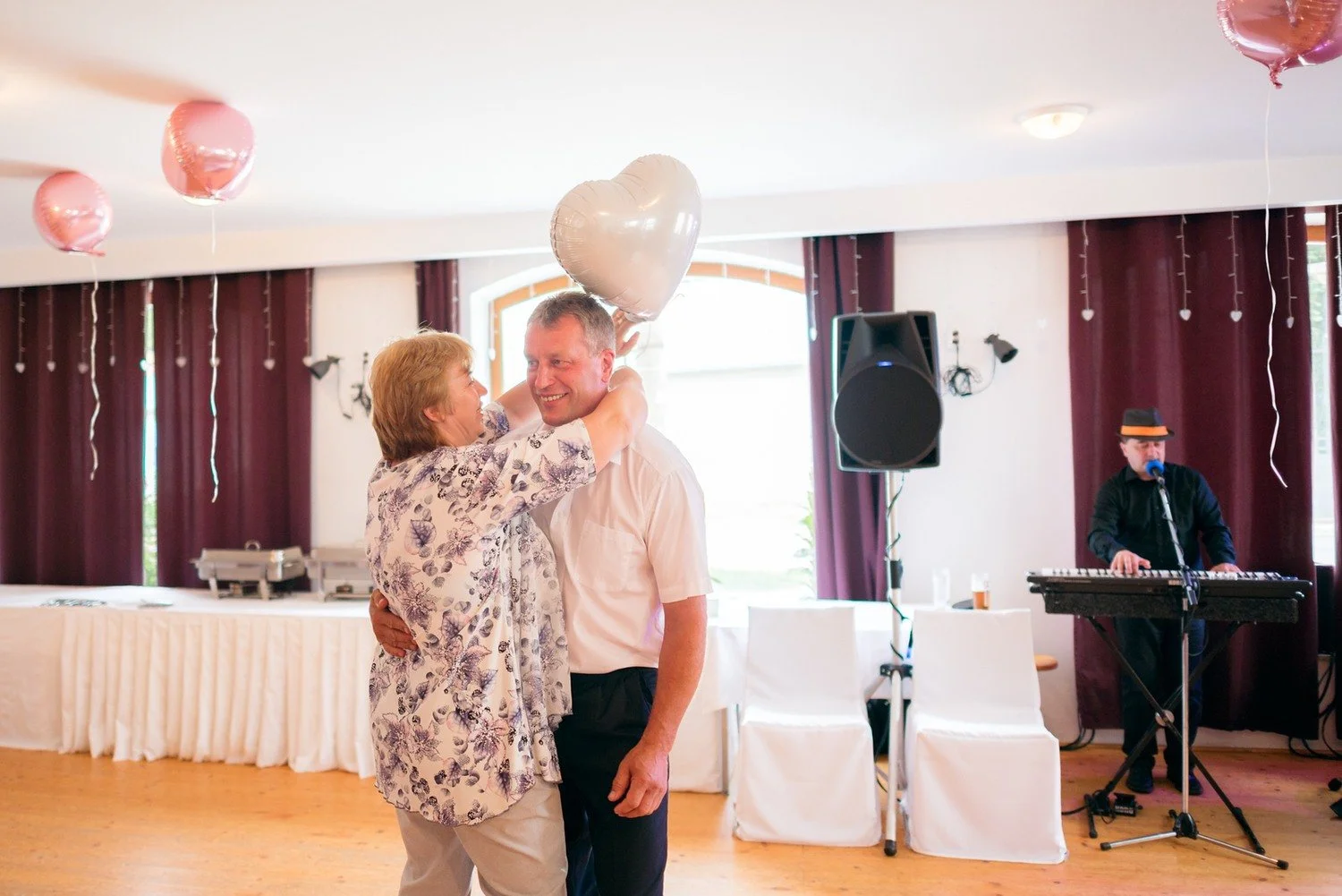 Žena líbá muže na tvář pod balonkem ve tvaru srdce, zatímco v pozadí hraje klávesista.