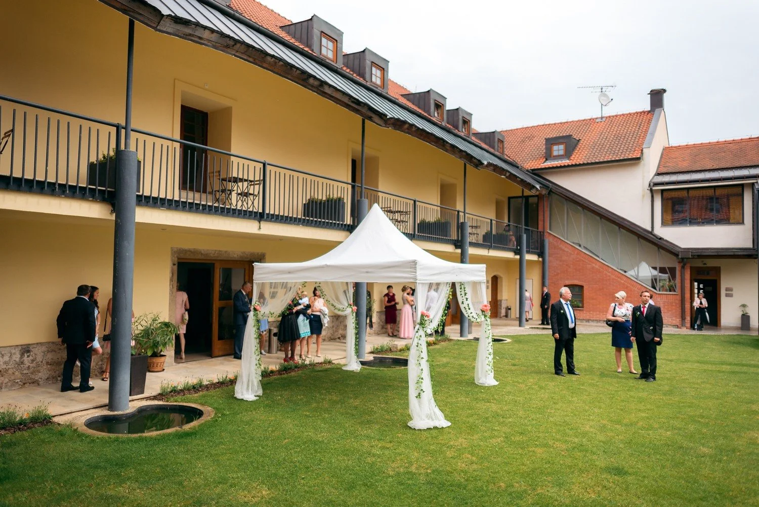 Na travnatém dvoře mezi budovami stojí bílý obřadní stan a několik čekajících hostů.