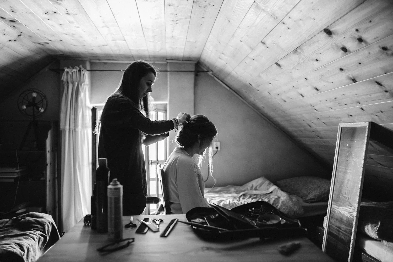Černobílý záběr úpravy účesu nevěsty v podkrovním pokoji se šikmým dřevěným stropem.