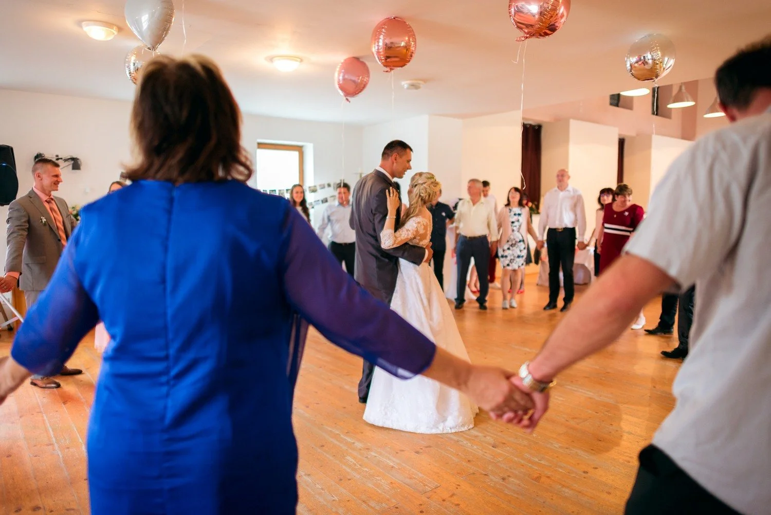 Hosté se drží za ruce v kruhu kolem tančících novomanželů pod růžovými a stříbrnými balonky.