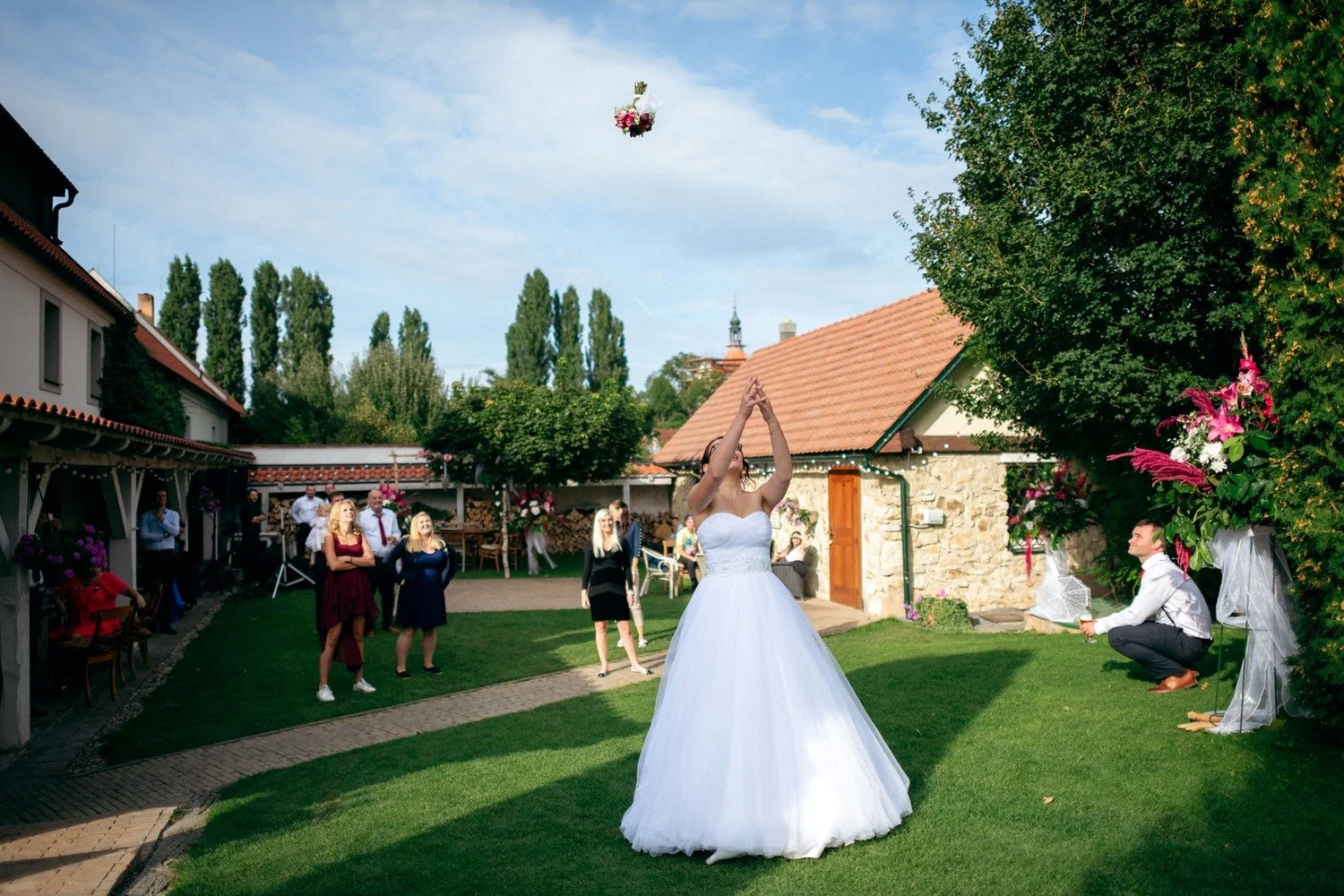 Žena v bílých šatech stojí zády se vzpaženýma rukama a kytice letí nad dvorem.