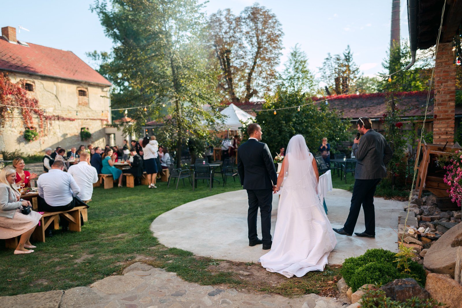 Dvojice přichází na kruhový parket mezi venkovními stoly a stojícími hosty.