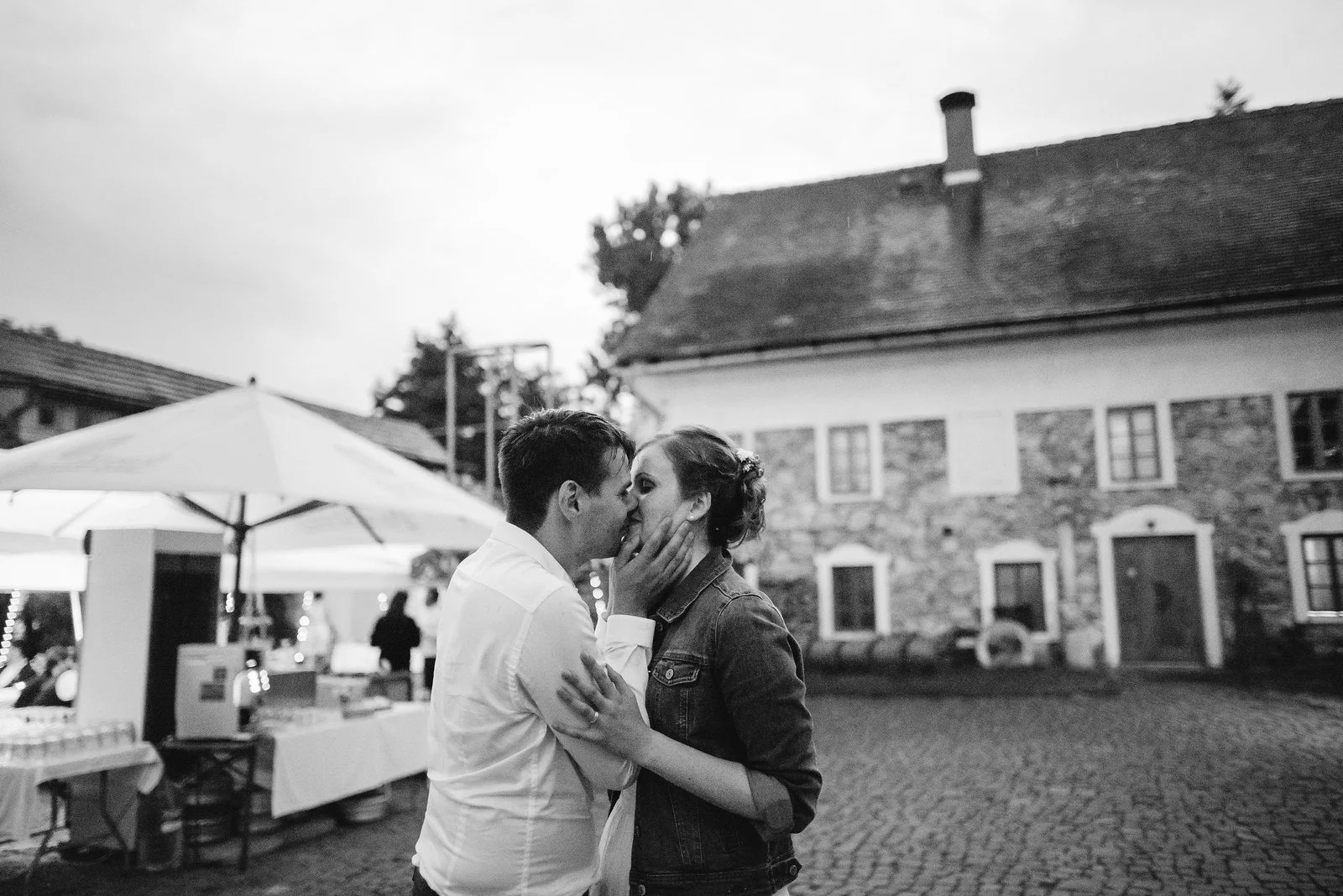 Dvě osoby se objímají na černobílém záběru před budovou a velkými bílými slunečníky.
