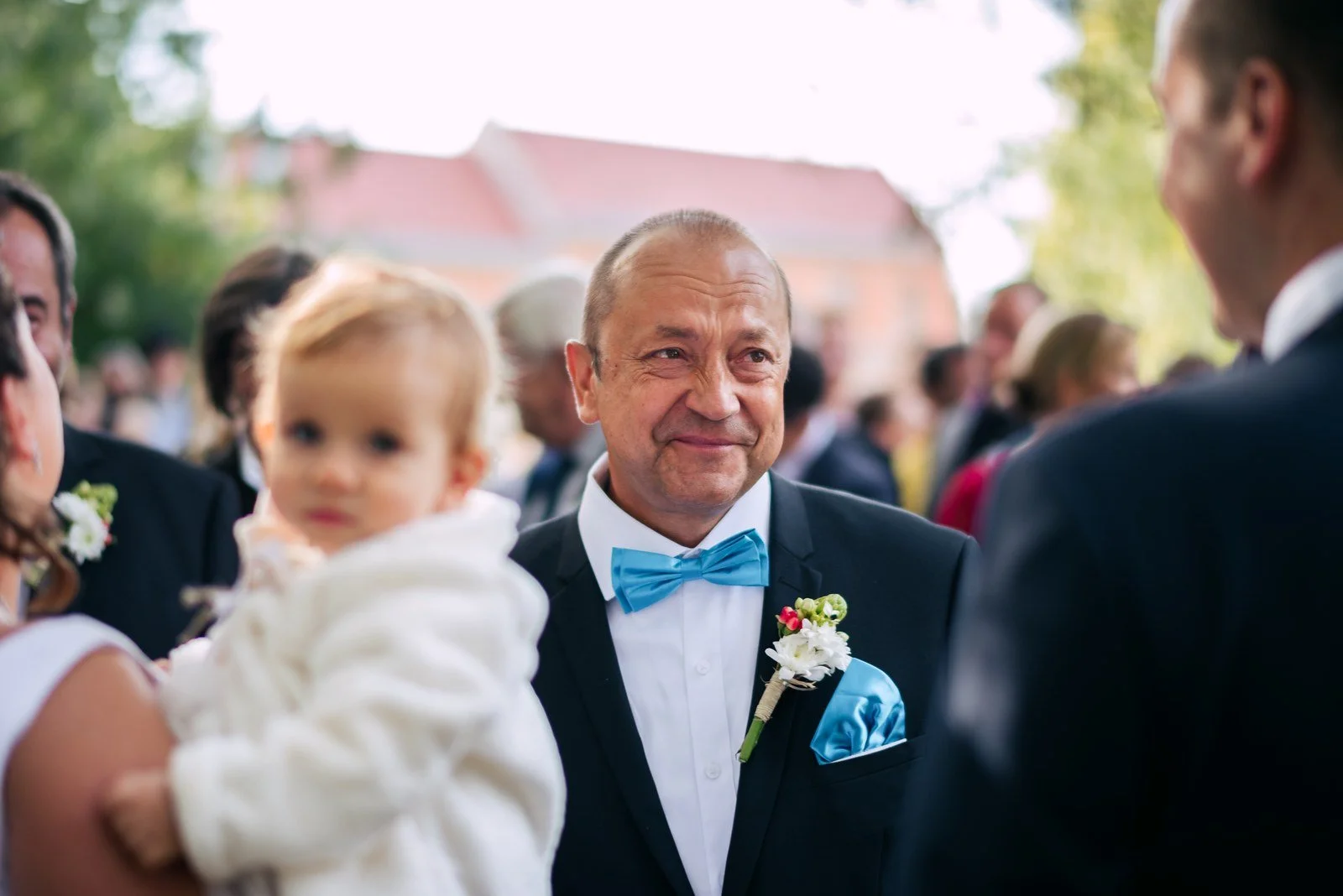 Muž s tyrkysovým motýlkem stojí mezi hosty, v popředí je malé dítě.