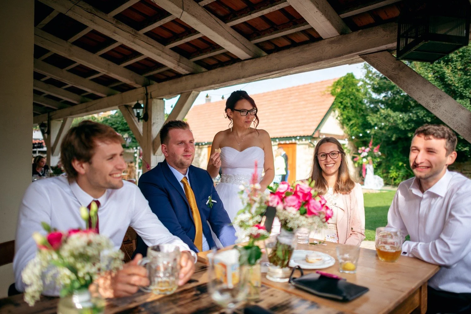 Pět slavnostně oblečených lidí sedí a stojí kolem stolu s květinami a sklenicemi.