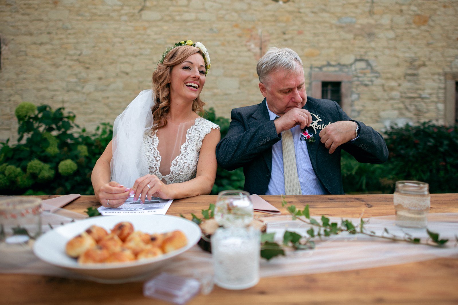Sedící žena v bílých šatech podává lžičku staršímu muži u venkovního stolu.