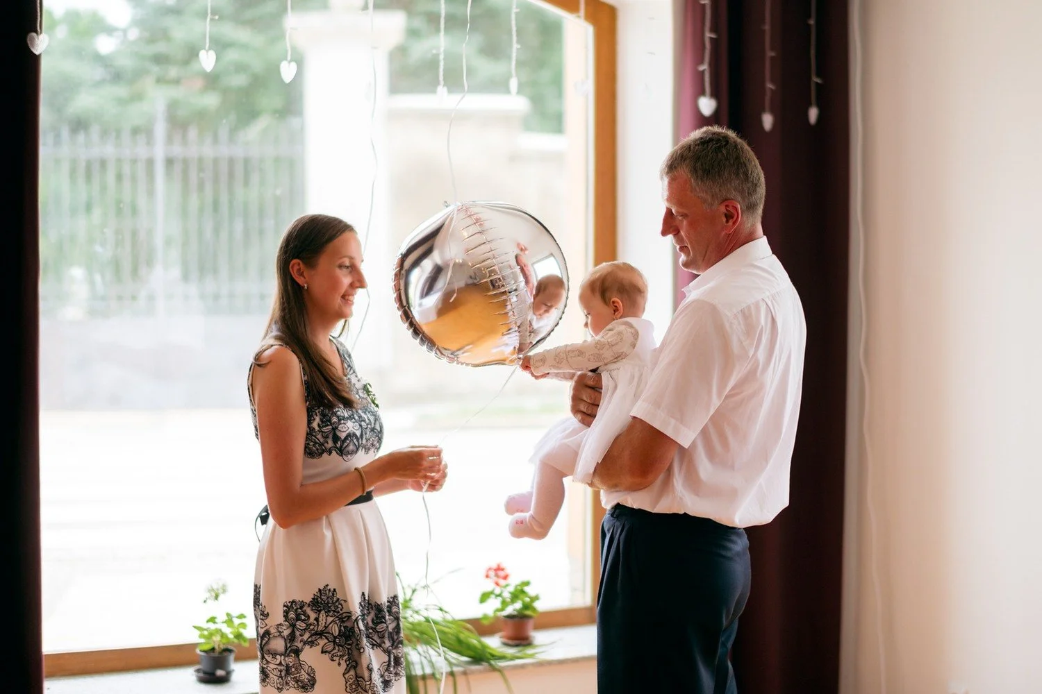 Muž drží malé dítě čelem ke kulatému stříbrnému balónku, zatímco vedle stojí žena se sklenkou.