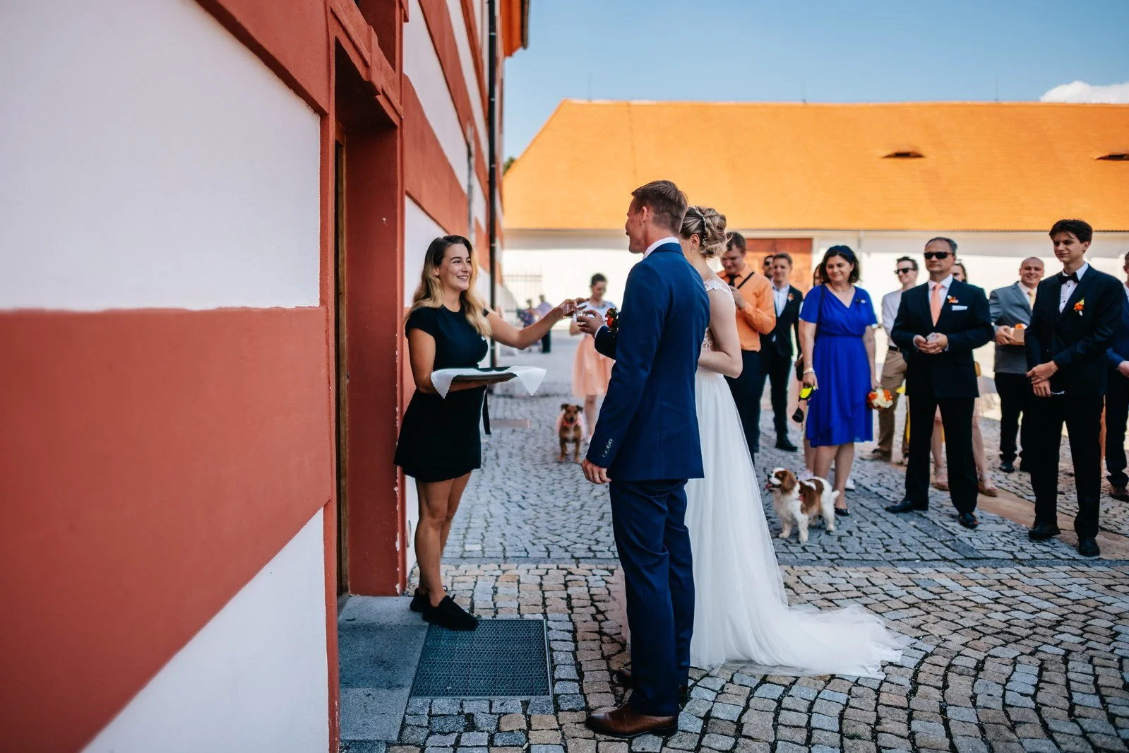 Novomanželé stojí na dlážděném nádvoří před skupinou hostů.