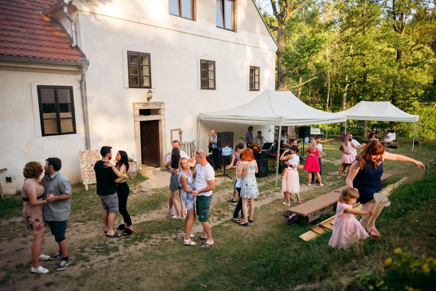 Hosté tančí venku mezi bílou budovou mlýna a stany v pozdním slunečním světle.